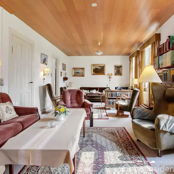 En stue med brune sofaer og en lænestol omgivet af hylder fyldt med bøger. Et sofabord med dug og blomster foran en tæppebelagt gulv, indrammet af billeder på væggene.