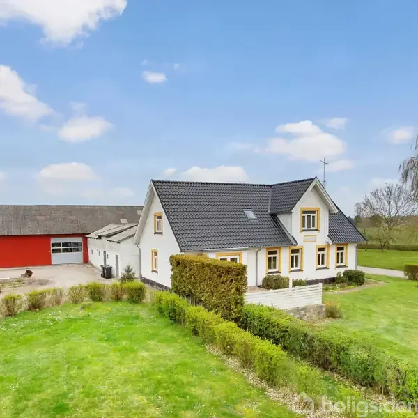 Hvidt hus med sort tag står stille ved siden af en rød lade; omgivet af grøn græsplæne og buske under en blå himmel med få skyer.