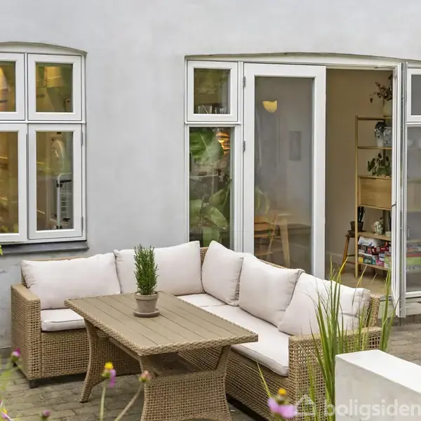 Fletsofa med hvide hynder står ved et træbord i en gårdhave foran en hvid bygning med åbne franske døre og vinduer, omkranset af blomster og grønne planter.