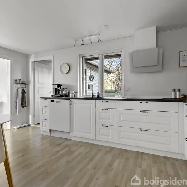 Køkken med hvide skabe og sorte bordplader, utrustet med køkkenapparater. En kaffemaskine og køkkenredskaber står på bordet. Et vindue giver udsigt til en have. Til højre ses en indbygget ovn.