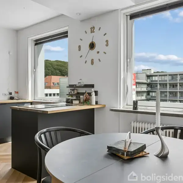 Køkken med sort bord og spisebord i forgrunden. Udsigt til bygninger gennem store vinduer. Der er en moderne væg-ur på væggen. Lysindfald fra venstre.