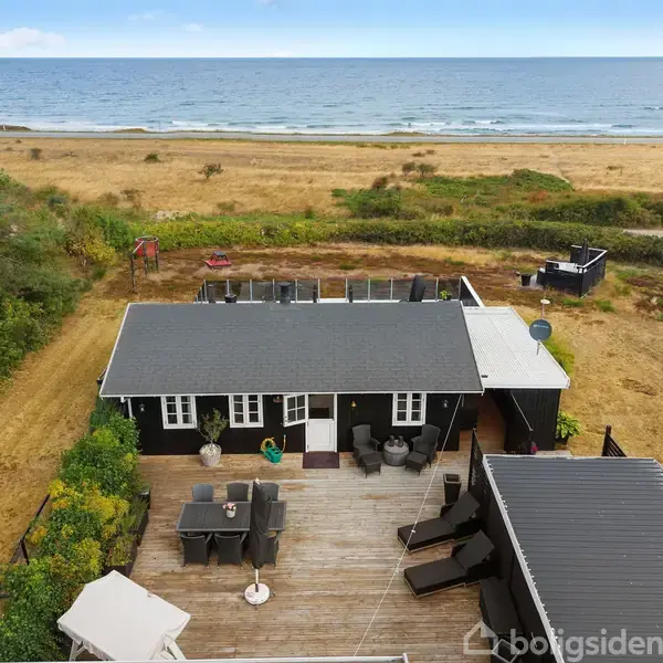Sommerhus med sort facade og gråt tag, står stille med møbleret terrasse i forgrunden. I baggrunden ses græsareal, træer og en sandstrand ved havet.