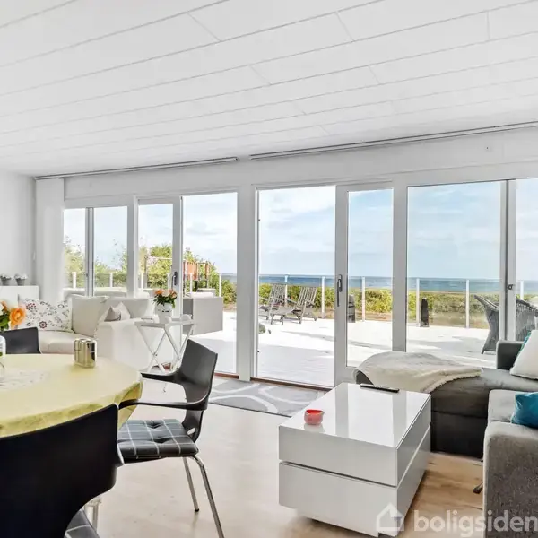 Stue med sofa og sofabord, vendt mod en stor skydedør med udsigt til terrasse og hav. Rundt spisebord med blomsterdekoration, tv på væggen.