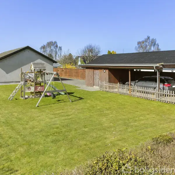 Hus med gyngestativ i haven, bil under carport. Omgivet af velplejet græsplæne og hegn. Klart blå himmel angiver solrige forhold.