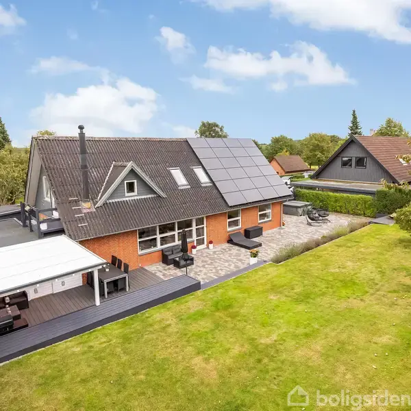 Et hus med solpaneler på taget står i en velholdt have. En terrasse med møbler er på bagsiden af huset. Omkringliggende træer og andre huse ses i baggrunden.