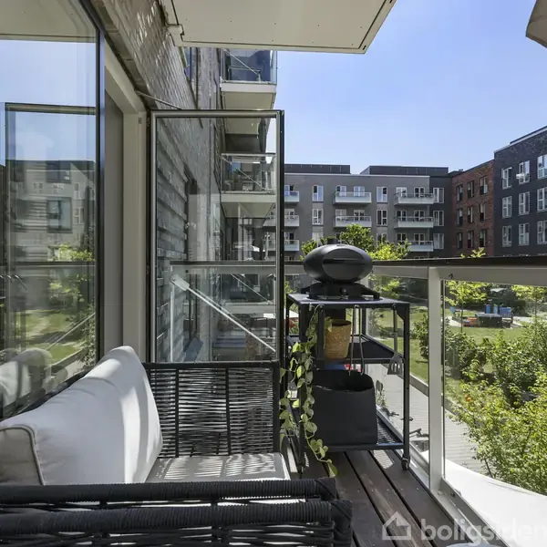 Altan med sort sofa med hvide hynder, en grill på stativ, og grønne planter. Omgivet af et moderne boligkompleks med grønt område i baggrunden under en klar blå himmel.