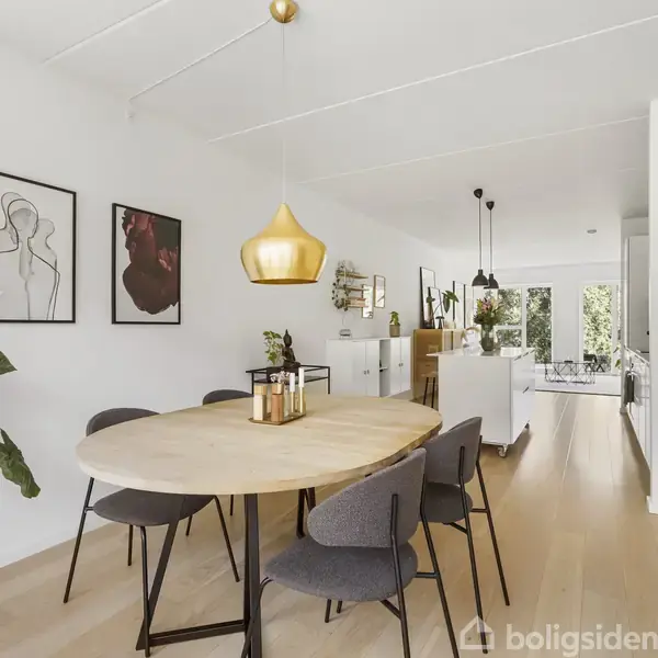 Et rundt spisebord med sorte stole omkring står midt i et lyst rum. En gylden lampe hænger over bordet. Væggene er dekoreret med moderne kunst, og der er en stor plante i hjørnet.