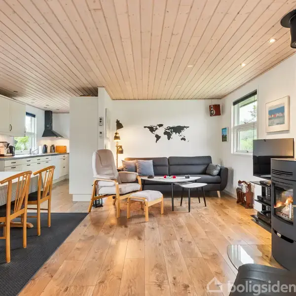 Et moderne, åbent stue-køkkenområde med en brændeovn i hjørnet. En sort sofa står ved en væg med et verdenskort, og spisebordet har fire stole. Trægulve og lys træloft.