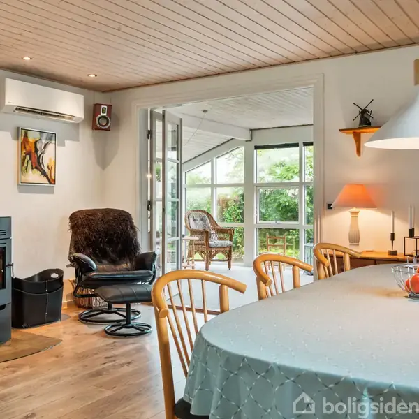 Brændeovn opvarmer en stue med spisebord, lampe og stol. Franske døre åbner ud til en veranda med grøn haveudsigt. Ingen tekst fundet.