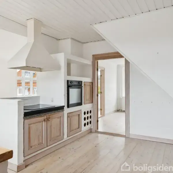 Et køkken med træskabe og indbygget ovn står mod en hvid væg. Der er en emhætte over en kogeplade, og rummet har lyse trægulve med en stor åbning til næste rum.