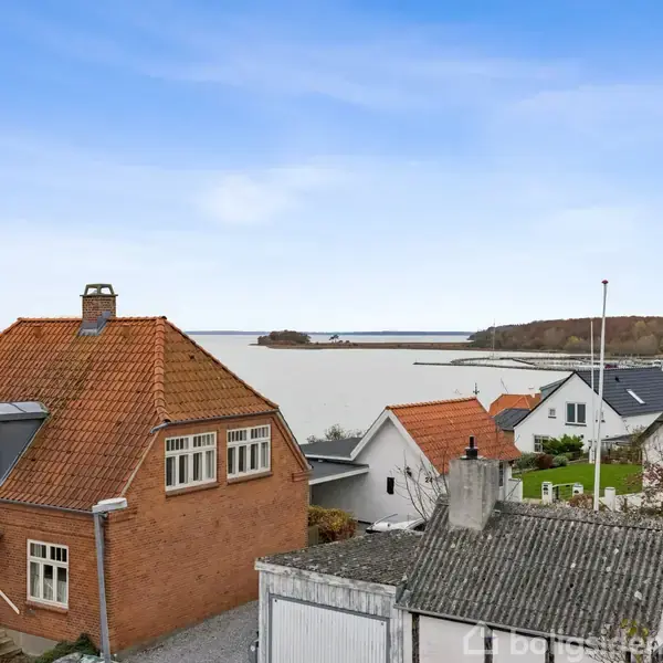 Et rødt murstenshus med skråt tag står i forgrunden, omgivet af mindre bygninger. I baggrunden ses en sø eller fjord, mens horisonten har skovklædte områder.