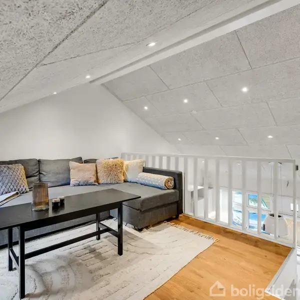 Sofa i gråt set-up står på et lyst tæppe ved et sofabord. Rummet er et loftsrum med skrålofter, hvide vægge og trælaminatgulv samt loftslys installeret.