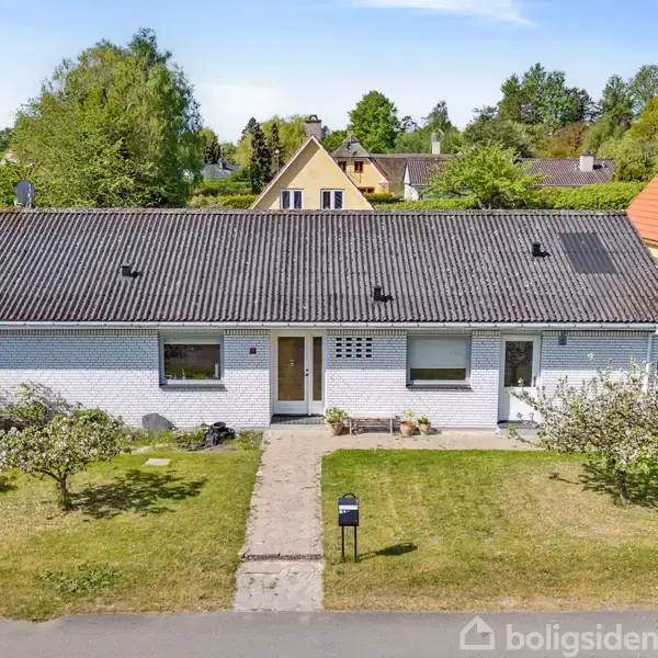 En hvid murstensvilla står midt i en velplejet have med stier, omgivet af grønne træer og nabohuse i et roligt villakvarter.