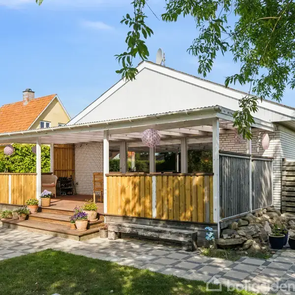 En én-etagers hvid træterrasse, overdækket med et tag, omgivet af havefliser, grønt græs og planter. Trærammen har potteplanter og møbler indenfor. Solskinsdag med blåt himmel.