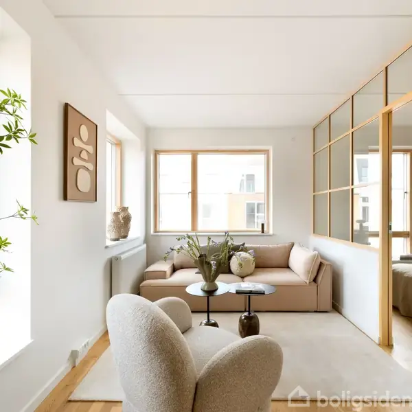 Stue med beige sofa og to lyse lænestole foran et rundt bord med blomster. Store vinduer og dekorativt kunstværk på væggen. Lyse trælameller deler rummet.