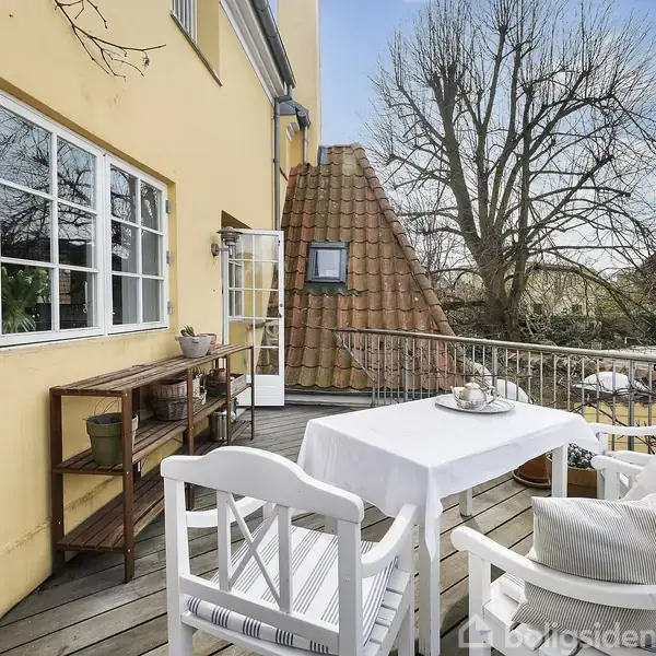 En terrasse med hvide havemøbler og bord med dug. Til venstre står en reol med planter. Baggrunden viser en gul væg, åbne døre, og et stort træ under en blå himmel.