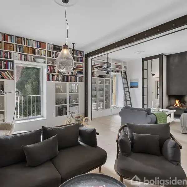 En stue med sorte sofaer er fyldt med reoler, der strækker sig til loftet med mange bøger. Et glasbord står foran sofaerne. Baggrunden viser en pejs og flere siddepladser.