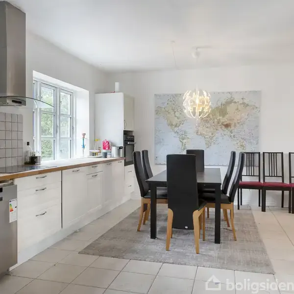 Køkkenet har et langt bord med sorte stole omkring. En lampe hænger over bordet. Bagved er et verdenskort på væggen. Køkkenbordet er mod venstre med hvide skabe.