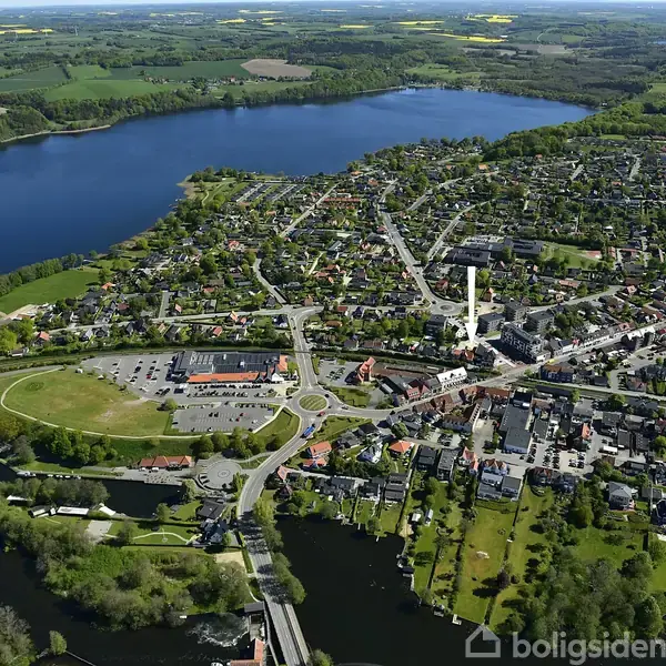 By med huse set fra oven, omgivet af floder og grønne marker. En stor sø er tæt på byen, mens veje og stier strækker sig gennem området.