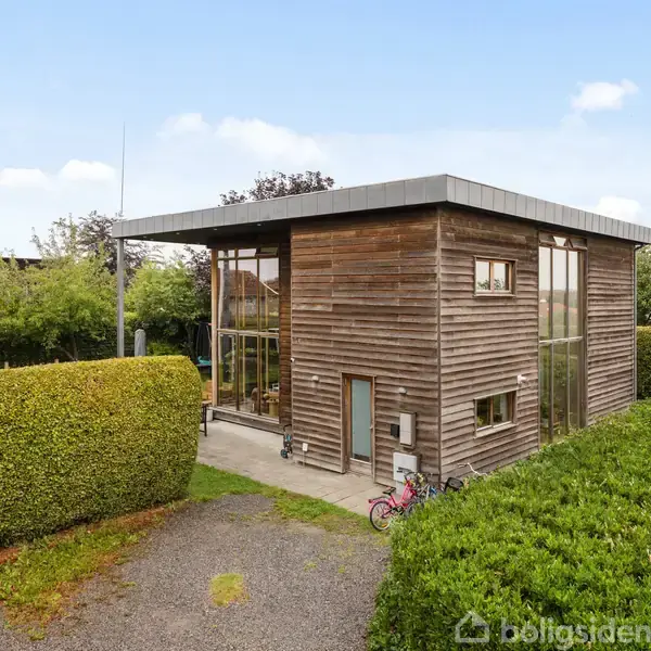 Et moderne træhus med store vinduer står stille omgivet af veltrimmede hække i en have. En cykel er parkeret ved siden af huset, hvilket tilføjer et hjemligt præg.