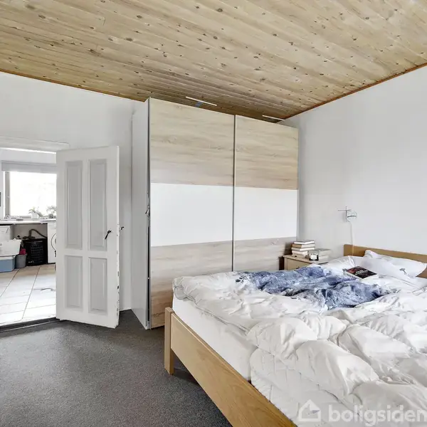 Et soveværelse med en dobbeltseng er uredt. Tøj ligger på sengen, og der er en stor skydedørsgarderobe. Døren fører til et køkken i baggrunden.