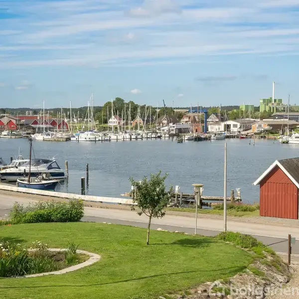 Både ligger i havnen ved kajen, omkranset af grønne områder, med rød-hvide træskure i nærheden og flere parkerede både langs vandet under en klar blå himmel.