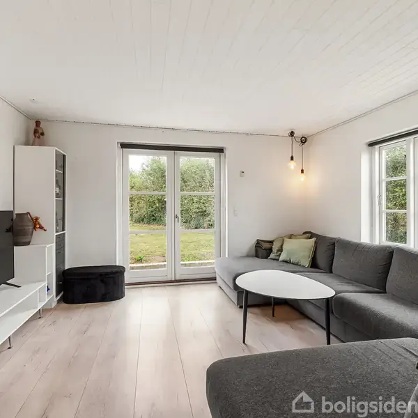 En stue med en grå hjørnesofa og et lille rundt bord. Sofaen står ved siden af store vinduer, som vender ud mod en have. Et fjernsyn er placeret på en hvid reol.