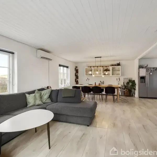 Grå sofa står i stue, omgivet af lyse trægulve og hvide vægge, vendende mod spisebord med sorte stole og moderne køkken. Store vinduer giver lys.