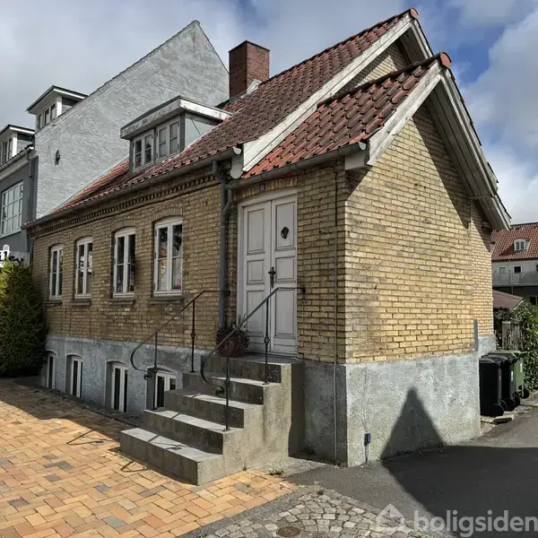 Et lille, gult murstenshus med rødt tegltag står ved en fortov. Trappen fører til en hvid hoveddør. Til venstre ses en café med skilt "smile".