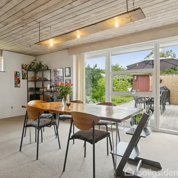 Et moderne spisebord af træ står i et lyst rum med glasdøre mod en terrasse. Stole omringer bordet, og planter samt kunst dekorerer væggene, skaber en indbydende atmosfære.