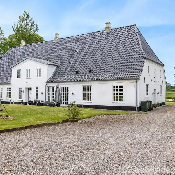 Et hvidt, traditionelt dansk landhus med mørkt tag står på en grusvej. Omkranset af grønne plæner og træer, under en lys blå himmel.