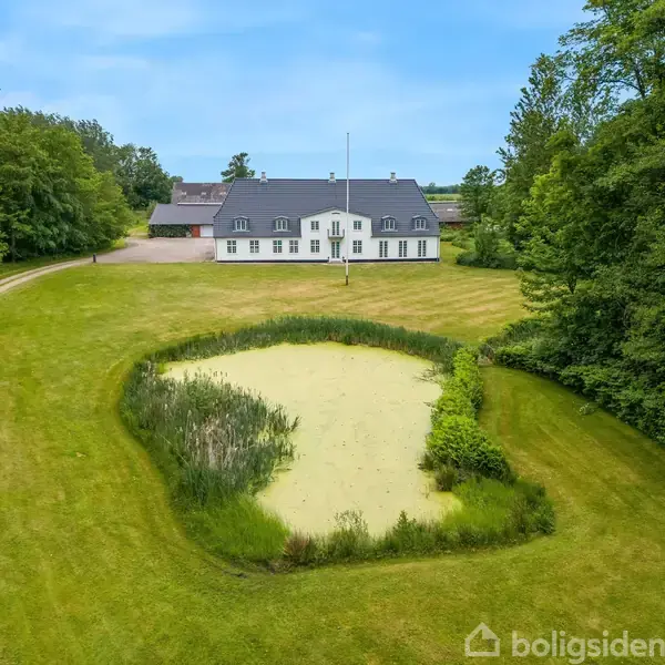 En stor, hvid bygning med sort tag står i midten af en græsplæne omkranset af træer. En lille, cirkulær dam ligger foran bygningen. Vejleder "Boligsiden" er synlig nederst.