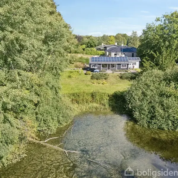 Et moderne hus med store vinduer står stille omgivet af frodig vegetation. Foran ses en lille sø, omgivet af træer og græs, som skaber en rolig atmosfære.