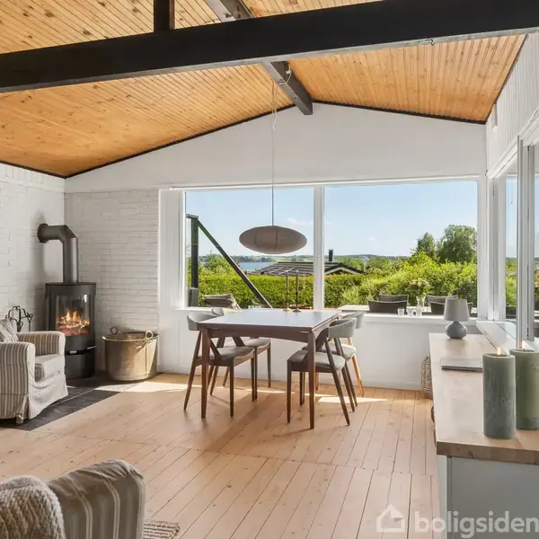En lys stue med trævægge og -loft har en brændeovn, et tæppe under en stol, og et spisebord med stole ved store vinduer, der vender ud mod naturen udenfor.