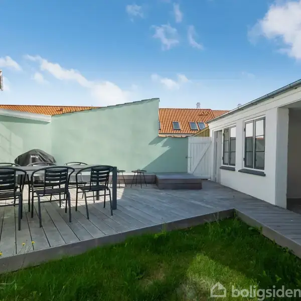 En terrasse har et sæt af borde og stole placeret på trægulvet. Omkring er der grønne græsarealer og en bygning med vinduer i baggrunden.