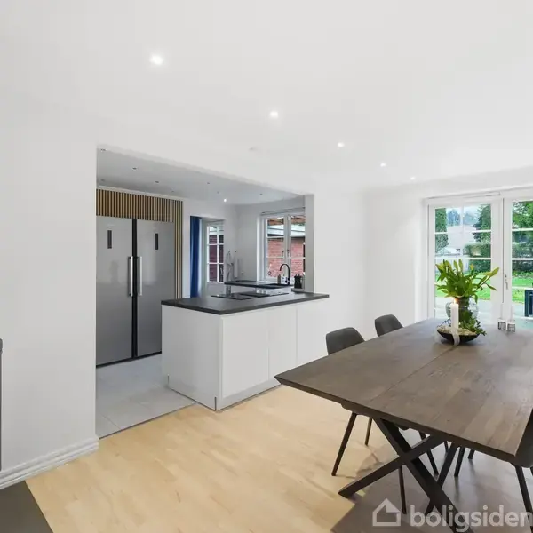 En brændeovn står i et moderne køkken-alrum. Et spisebord med stole ses ved siden af glasdøre, der fører ud til haven. Rum indeholder også et køleskab og køkkenø.