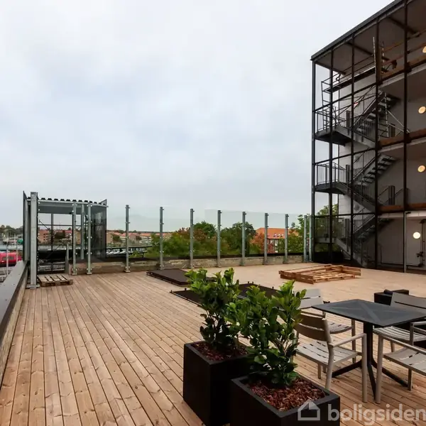 Stor tagterrasse med trædæk, hvor borde og stole er placeret. Der er planter i krukker, og i baggrunden ses grønne områder og en togbane.