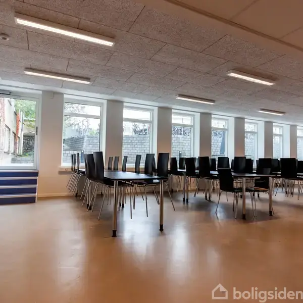 Stole står stablet på borde i et lyst lokale med store vinduer, der giver udsigt til en udendørs mur. En lille trappe fører op til en dør på venstre side.