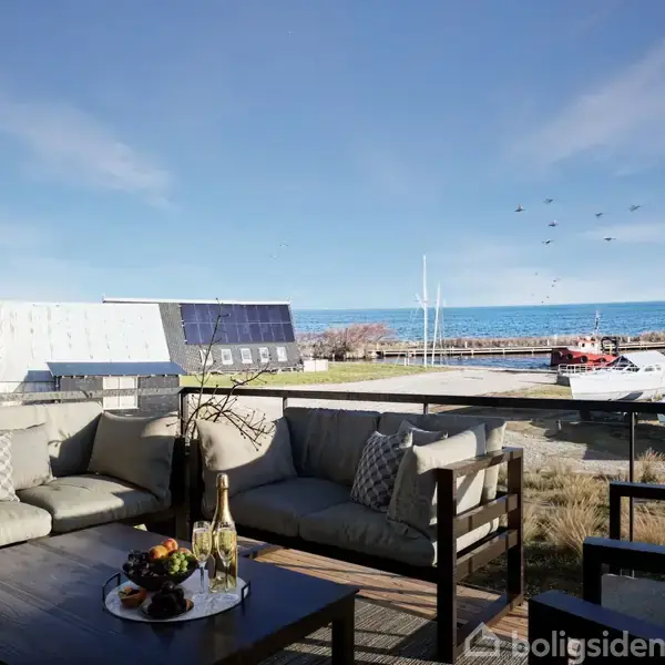 Sofagruppe står på terrasse med havudsigt. Bordet er pyntet med en bakke, champagne og frugter. Både ses ved havnen, og himlen er blå med nogle fugle.