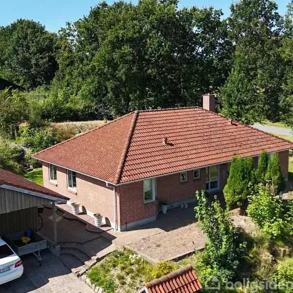 En murstensvilla med rødt tegltag står ved siden af en carport med en hvid bil under. Omgivet af grønne buske og træer i et stille kvarter.