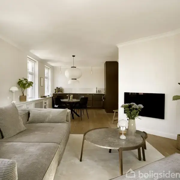 En lys stue med sofa og sofabord. I baggrunden ses et indrettet køkken med spisebord og hængende lampe. Grønne planter tilføjer et friskt udtryk.