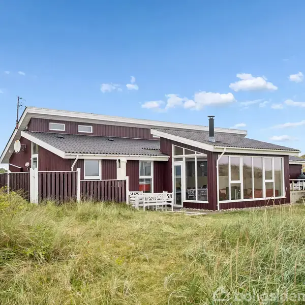 Et rødt træhus med store vinduer står midt i et græsbeklædt klitområde under en blå himmel med spredte skyer. Huset har en terrasse med hvidt havemøblement.