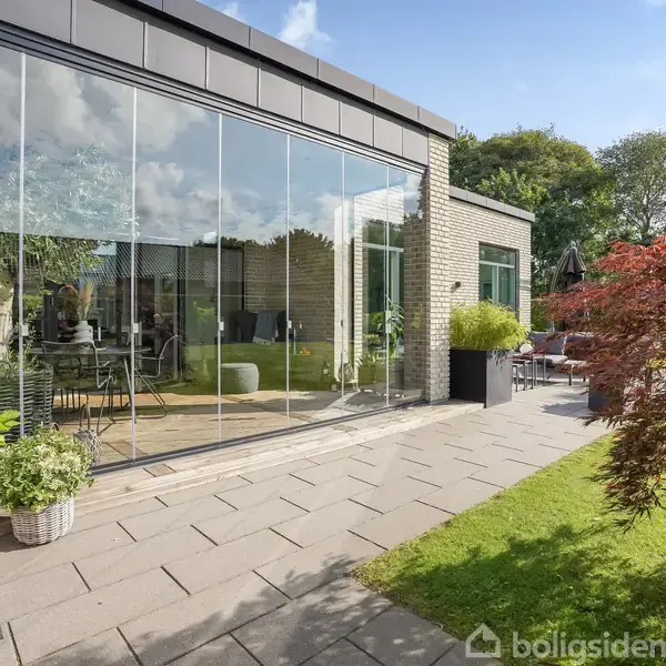 En moderne glasfacade reflekterer haven med grøn vegetation og en terrasse med flisebelægning. Sollys skaber et lyst udtryk, med planter i potter ved bygningen.