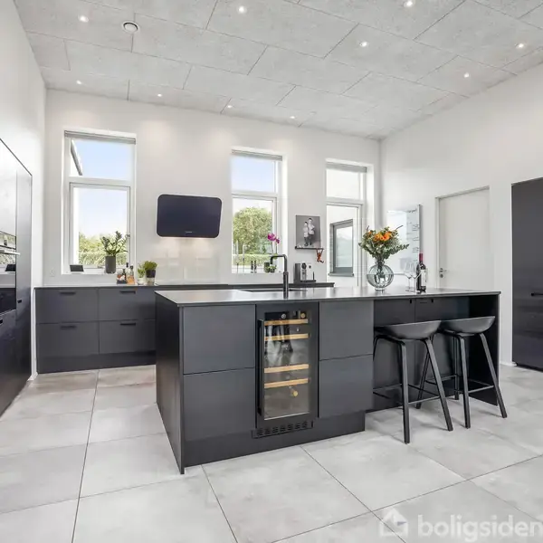 Et moderne køkken med mørke skabe og en central ø, som indeholder en vinkøler. To sorte barstole står ved øen. Rummet har store vinduer, der giver naturligt lys.