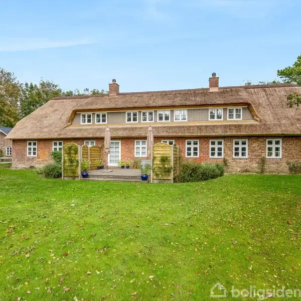 Hus med stråtag og mange vinduer står i en have med grøn græsplæne og træer. Huset har en murstensfacade og en trædør, omgivet af grønne planter og blå krukker.