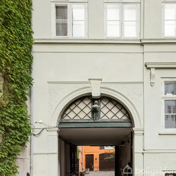 En hvid murstensbygning med en stor bueformet portåbning. Indgraverede årstal "1751 & 1783" ses over porten. Bygningens facade har grøn beplantning til venstre og vinduer omkring.