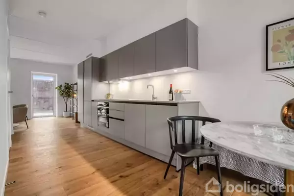 Moderne køkken med grå skabe og indbygget ovn opstillet mod en hvid væg. Rundt marmorbord med sort stol i forgrunden. Lyst trægulv og en dør til en terrasse i baggrunden.