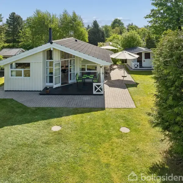 Et hvidt sommerhus med sorte detaljer står på en velplejet græsplæne. Huset har en terrasse med møbler og en parasol, omgivet af frodig, grøn vegetation.