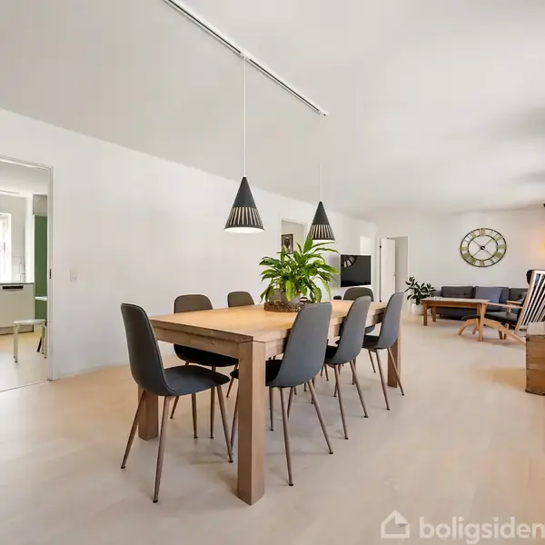 Spisebord med sorte stole står på lyst trægulv, under moderne lamper. Til venstre ses køkkenindgang, mens en lys stue med sofa og vægur er til højre.