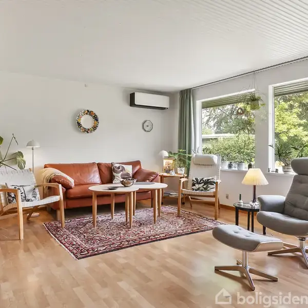 En stue med en brun lædersofa, sofabord, og en grå lænestol. Rummet har trælaminatgulv, grønne planter og store vinduer, der viser en udsigt til en have udenfor.
